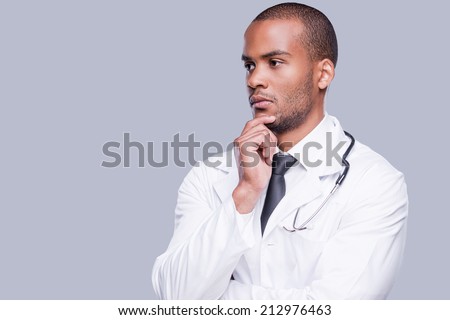 Thoughtful doctor. Thoughtful African doctor holding hand on chin and looking away while standing against grey background