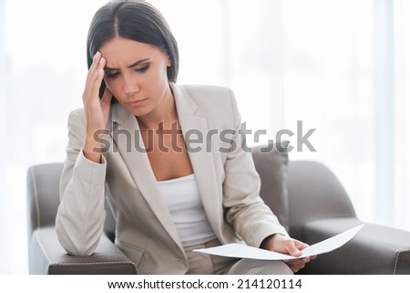 Tired of paperwork. Depressed young businesswoman in suit holding document and touching head with hand while sitting at the chair
