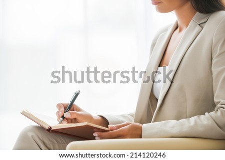 Making business notes. Side view of young businesswoman in suit writing something in note pad while sitting at the chair