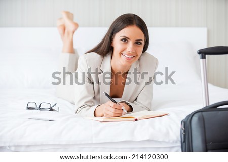 Making some urgent notes. Beautiful young businesswoman in suit writing something in her note pad and smiling while lying in the bed at the hotel room