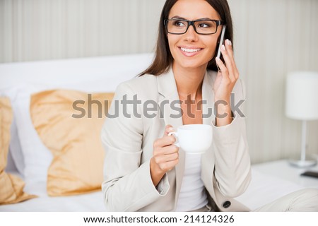 Taking time to talk with nearest. Beautiful young smiling businesswoman in suit drinking coffee and talking on mobile phone while sitting on the bed in hotel room