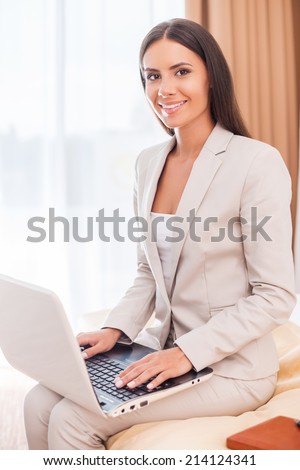 I love my job! Beautiful young businesswoman in suit working on laptop and smiling while sitting on the bed