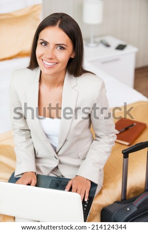 Staying in touch with my colleagues. Top view of beautiful young businesswoman in suit working on laptop and smiling while sitting on the bed in hotel room