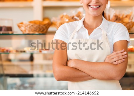Happy baker. Close-up of beautiful young woman in apron keeping arms crossed while standing in bakery shop