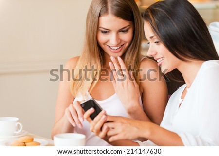Can you believe it? Two attractive young women looking at mobile phone and smiling while sitting in coffee shop together
