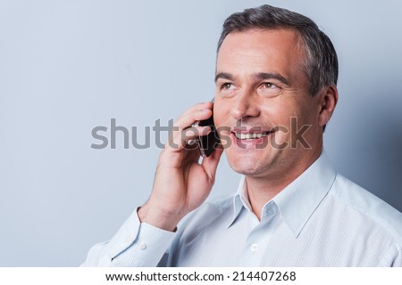 Good talk. Portrait of confident mature man in shirt talking on the mobile phone and smiling while standing against grey background