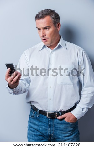 Shocking news. Surprised mature man in shirt holding mobile phone and looking at it while standing against grey background