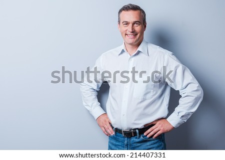Confident and successful. Confident mature man in shirt looking at camera and smiling while holding hands on hip and standing against grey background