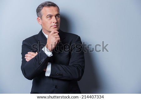 Thinking about solutions. Thoughtful mature man in formalwear holding hand on chin and looking away while standing against grey background