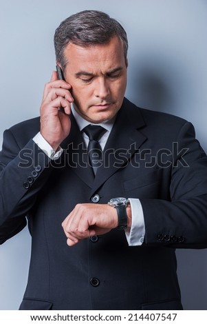 He knows that time is money. Confident mature man in formalwear checking the time and talking on the mobile phone while standing against grey background
