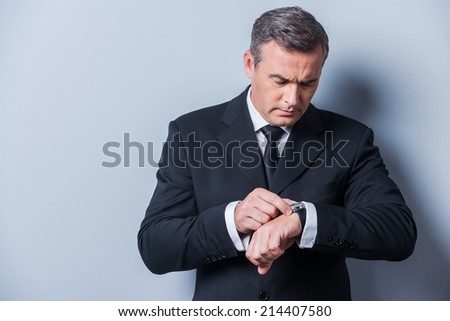 Businessman checking the time. Confident mature man in formalwear checking the time while looking at his watch and standing against grey background