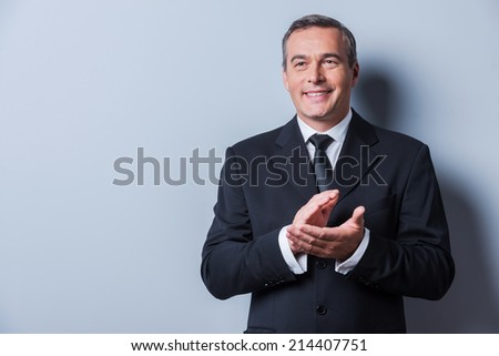 Celebrating success. Confident mature man in formalwear clapping hands and smiling while standing against grey background