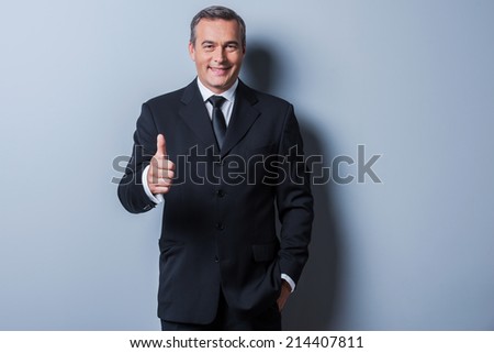 Thumb up for success! Cheerful mature man in formalwear showing his thumb up and smiling while standing against grey background