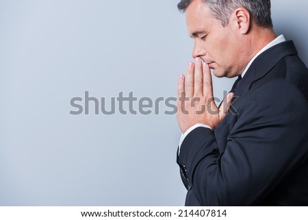Praying for success. Side view of concentrated mature man in formalwear holding hands clasped near face and keeping eyes closed while standing against grey background