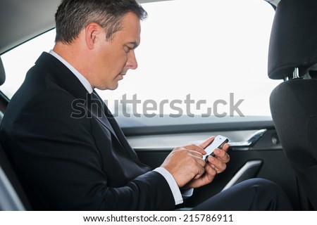 Sending an urgent message. Side view of confident mature businessman typing message on his smart phone while sitting on the back seat of a car