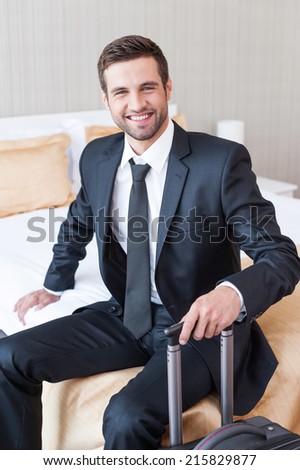 Executive on the Go. Confident young businessman in formalwear carrying suitcase and smiling while sitting on the bed in hotel room