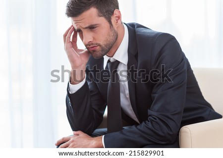 Depressed businessman. Side view of depressed young businessman in suit touching head with hand while sitting at the chair