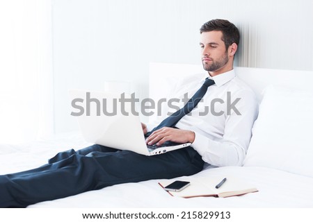 No time to relax. Confident young man in shirt and tie working on laptop while lying in bed at the hotel room