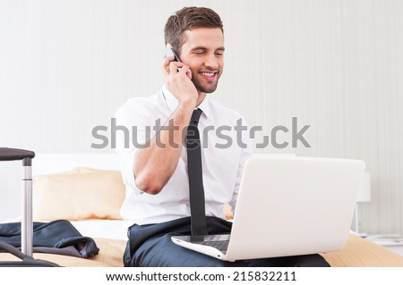Staying in touch with colleagues. Handsome young man in shirt and tie working on laptop and talking on the mobile phone while sitting on the bed in hotel room