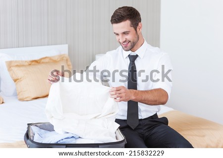 Choosing new shirt to wear. Handsome young man in shirt and tie unpacking his suitcase and smiling while sitting on the bed in hotel room