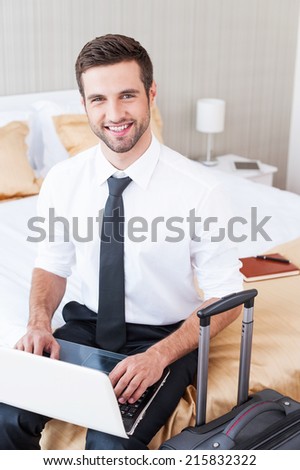 Staying in touch anytime and everywhere. Top view of handsome young man in shirt and tie working on laptop and smiling while sitting on the bed in hotel room