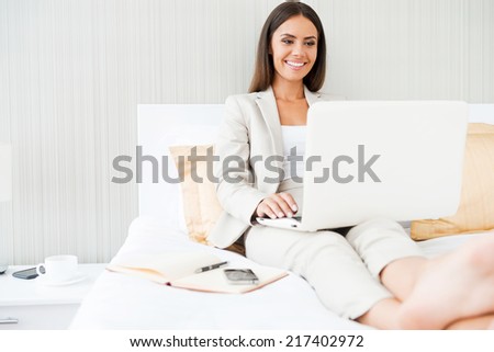 Working in hotel room. Confident young businesswoman in suit working on laptop and smiling while sitting in bed at the hotel room