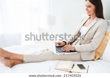 Businesswoman in hotel room. Beautiful young businesswoman in suit working on laptop and smiling while sitting in bed at the hotel room