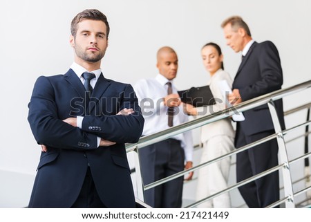 Confident businessman. Low angle view of confident young man in formalwear keeping arms crossed and looking at camera while standing at the staircase with people in the background