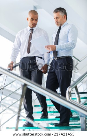 Discussing business contract. Two confident businessmen discussing something and looking at paper while moving down by staircase