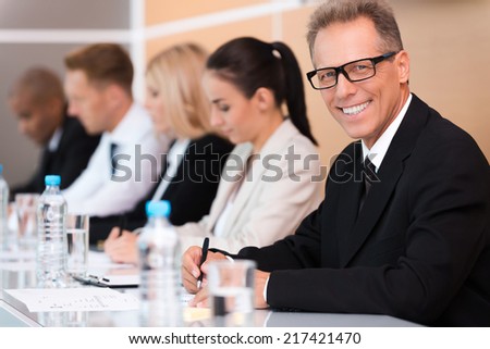 Business conference. Group of business people sitting in a row and writing something in their note pads while confident mature man in formalwear looking at camera and smiling