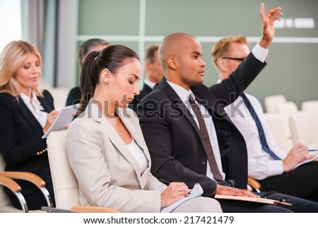 I have a question! Group of business people in formalwear sitting at the chairs in conference hall and writing something gin their note pads while handsome African man raising his arm