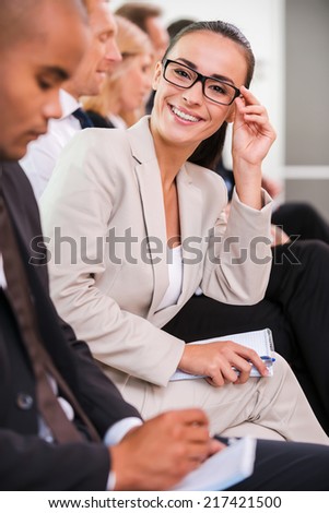 Confident business expert. Side view of business people sitting in a row and writing something in their note pads while confident young woman adjusting her eyeglasses and smiling