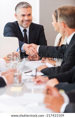 Welcome on board! Confident mature man in formalwear shaking hand to one of his colleagues and smiling while sitting at the table together