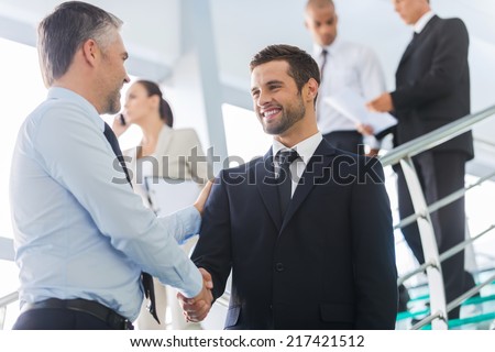 Businessmen shaking hands. Two confident businessmen shaking hands and smiling while standing at the staircase together with people in the background