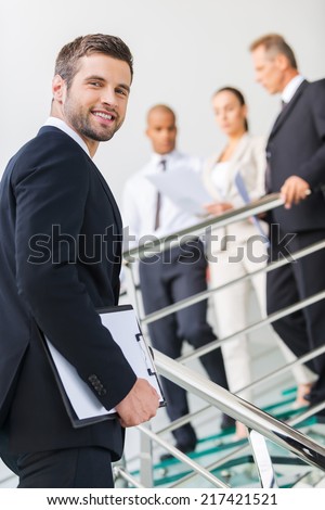 Young and successful. Low angle view of confident young man in formalwear looking over shoulder and smiling while moving up by staircase with people in the background