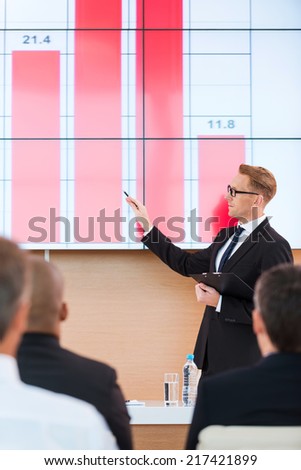 Presentation in conference hall. Confident young man in formalwear pointing projection screen with graph on it while making presentation in conference hall with people on the foreground