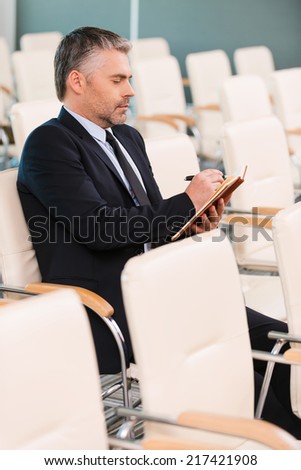 The one and only. Side view of concentrated mature man in formalwear writing something in note pad while sitting on the chair in empty conference hall