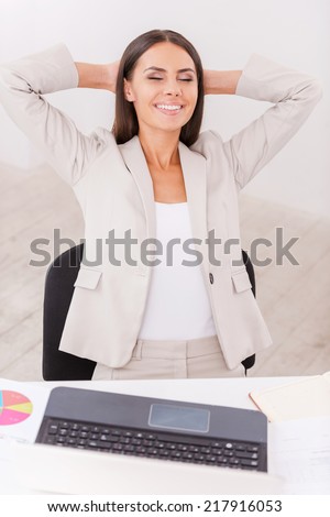 Satisfied with a job done. Happy young businesswoman in suit holding hands behind head and smiling while sitting at her working place