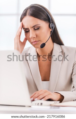 Sick and tired. Depressed young businesswoman in headset touching her head with hand and keeping eyes closed while sitting at her working place