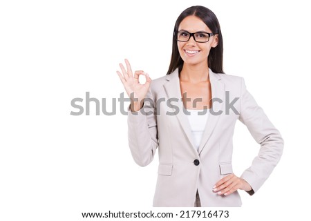 Everything is OK. Beautiful young businesswoman in suit gesturing OK sign and smiling while standing against white background