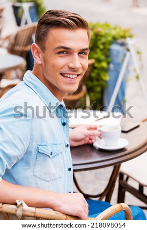 Spending good time in cafe. Rear view of cheerful young man drinking coffee and looking over shoulder while sitting in sidewalk cafe