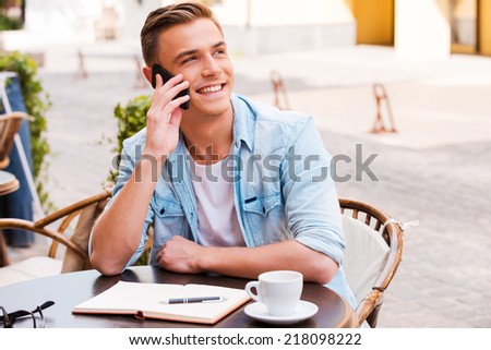 Talking with friends. Confident young man talking on the mobile phone and smiling while sitting in sidewalk cafe