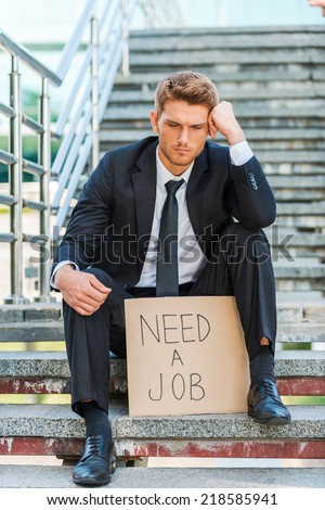 Need a job. Depressed young man in formalwear holding poster with job text message while sitting on the staircase