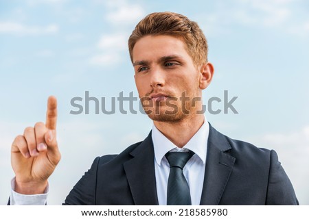 Working on transparent wipe board. Confident young man in formalwear touching a transparent wipe board while standing against sky background