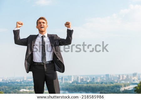 Business winner. Happy young man in formalwear keeping arms raised and expressing positivity while standing outdoors with cityscape in the background