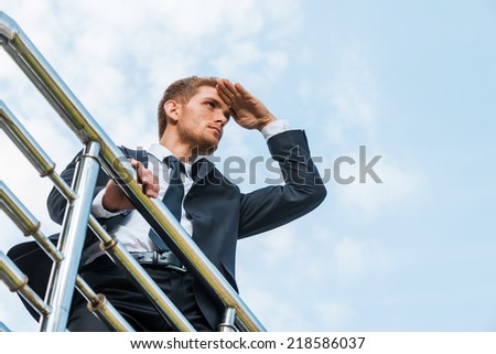 Looking for new opportunities. Low angle view of confident young man in formalwear holding hand on forehead and looking away while standing outdoors and leaning at the metal railing