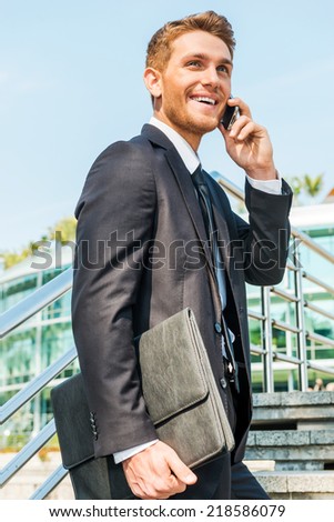 Great news! Low angle view of confident young man in formalwear talking on the mobile phone and smiling while moving up by staircase