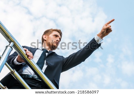 He knows the right direction. Low angle view of confident young man in formalwear pointing away while standing outdoors and leaning at the metal railing
