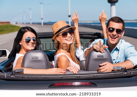 Friends in convertible. Group of young happy people enjoying road trip in their convertible while three of them looking over shoulder and smiling