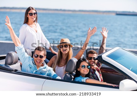 Great day for a ride. Group of young happy people enjoying road trip in their white convertible and raising their arms